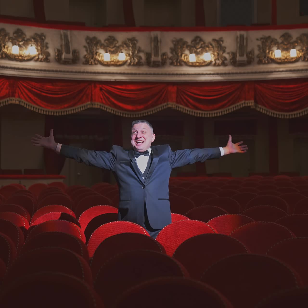 Happy rich man in empty theater hall. Successful businessman in a suit standing alone among empty chairs in theater and waving his hands.