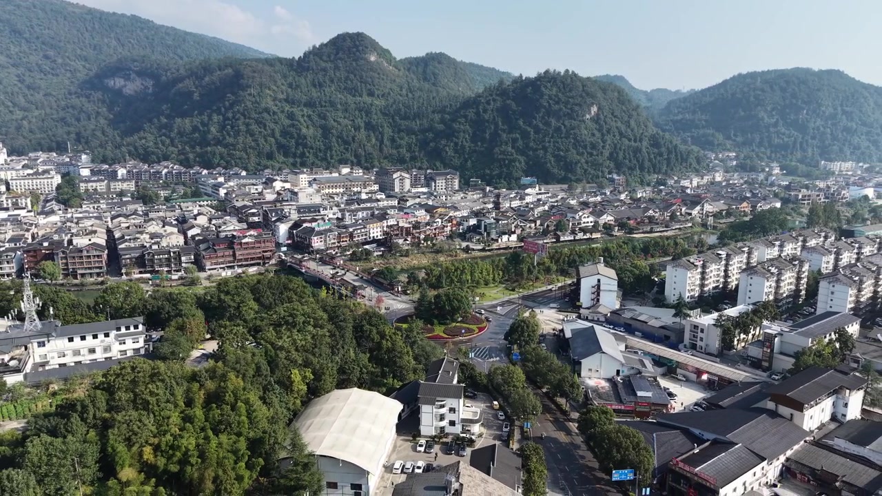 Lenta panoramica aerea verso destra su una valle con edifici bassi e palazzine, colline boscose e montagne carsiche sullo sfondo, cielo nuvoloso, Zhangjiajie, Hunan, Cina