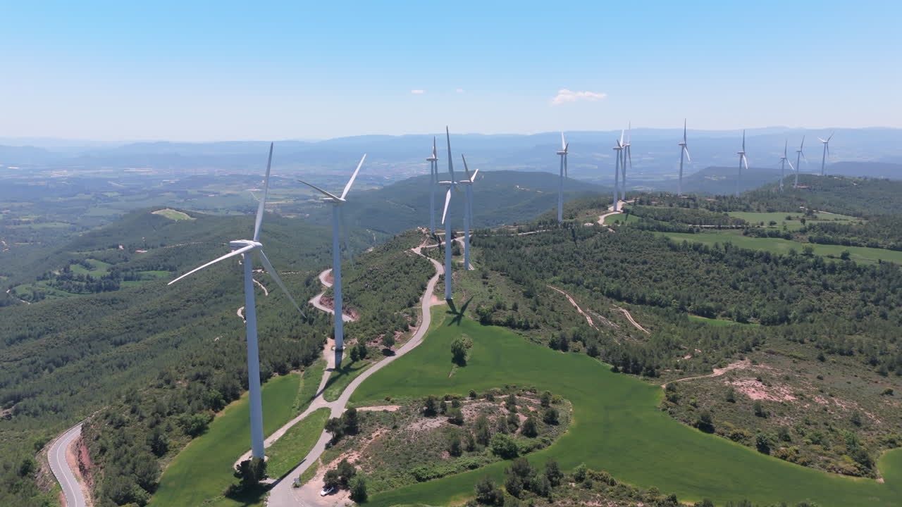 Drone shot panning slowly across a wind farm on mountain ridges, with tall wind turbines spinning under a clear blue sky and surrounded by green forest and farmland