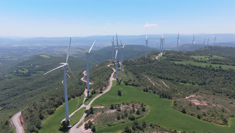 drone shot panning slowly across a wind farm on mountain ridges, with tall wind turbines spinning under a clear blue sky and surrounded by green forest and farmland