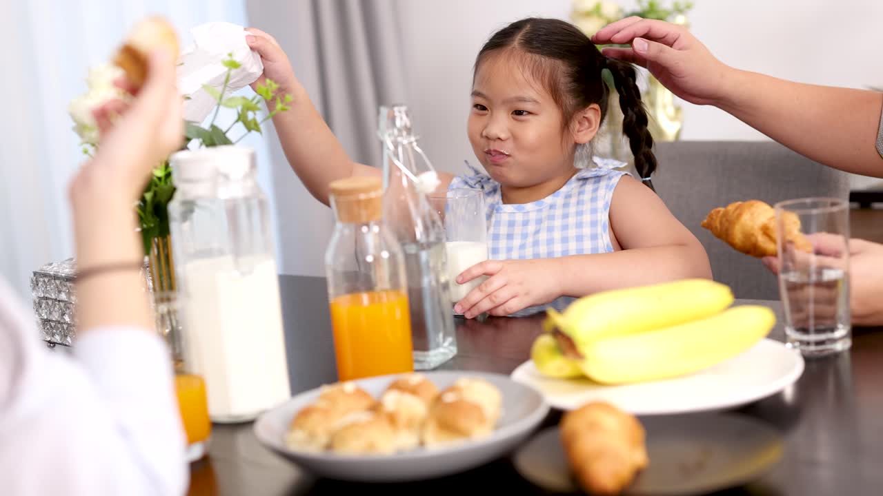 Mother helps young Asian girl wipe mouth at breakfast table in bright, cozy kitchen