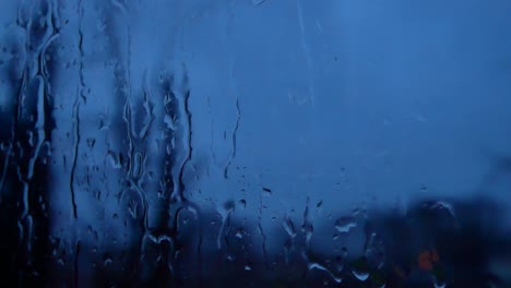 Rainy-storm-droplets-at-night,-moody-weather-fall-down-window-glass-closeup-bokeh