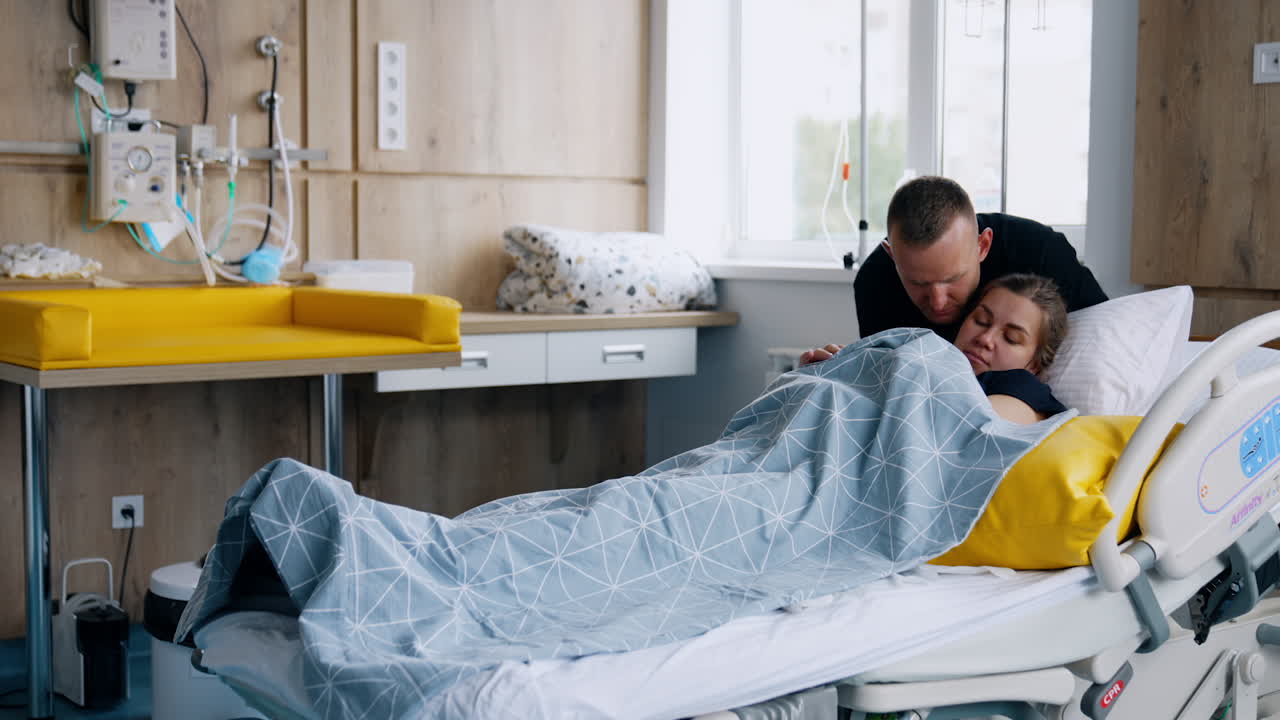 First time parents in the after labor ward. Woman lies in bed feeding a newborn and man stands near her.