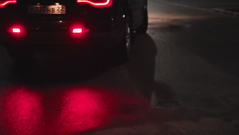 close-up view of a black car reversing at night, its back light reflecting on the ground. a white car approaches in the background