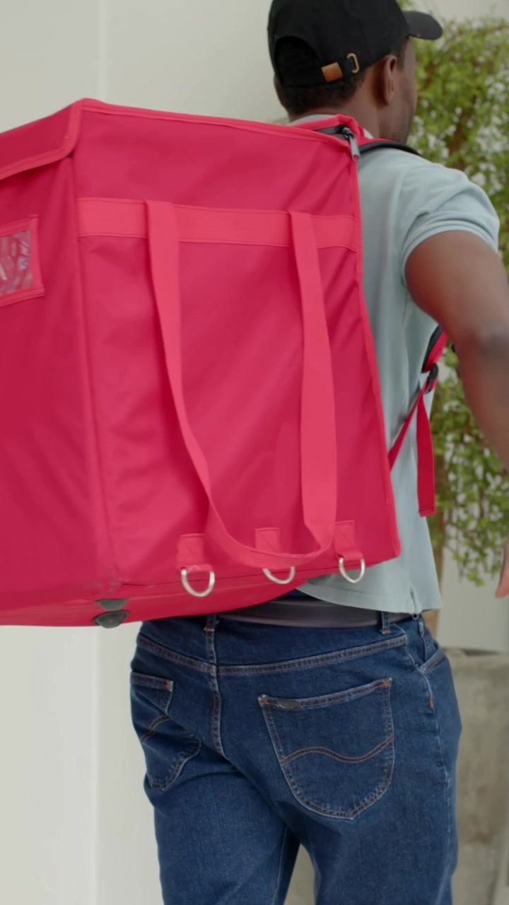 Video vertical: Repartidor con polo ajustando una bolsa roja en la puerta después de apoyarse en su scooter, para una entrega.