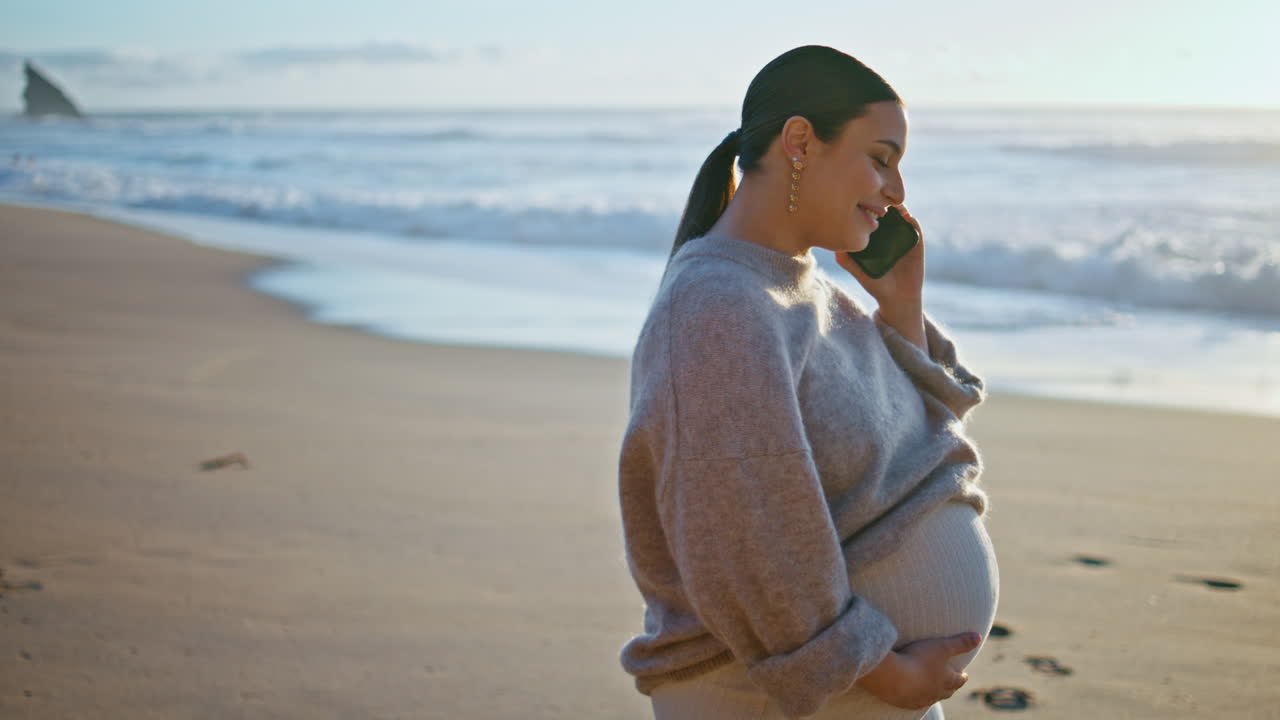 미래의 어머니는 스마트폰으로 이야기하고 해변을 가까이에서 고 있습니다. 임신한 여성이 전화하고 있습니다.