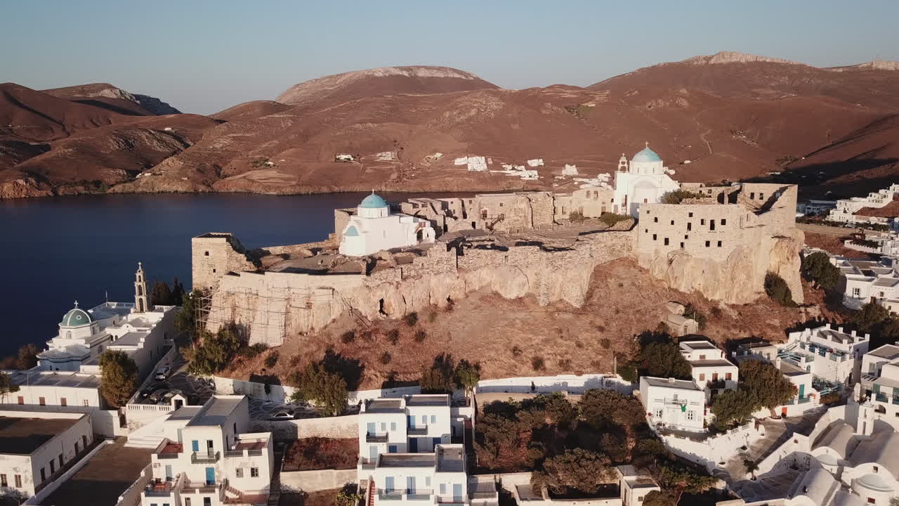 Greece, Astypalea Castle drone footage from a perfect angle,camera moves from left to right slowly.Two white chapels with blue domes are shinning early in the morning.Castle surrounded by white houses