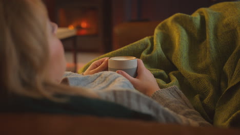 Close-Up-Of-Woman-At-Home-In-Lounge-Lying-On-Sofa-Looking-At-Cosy-Fire-With-Hot-Drink