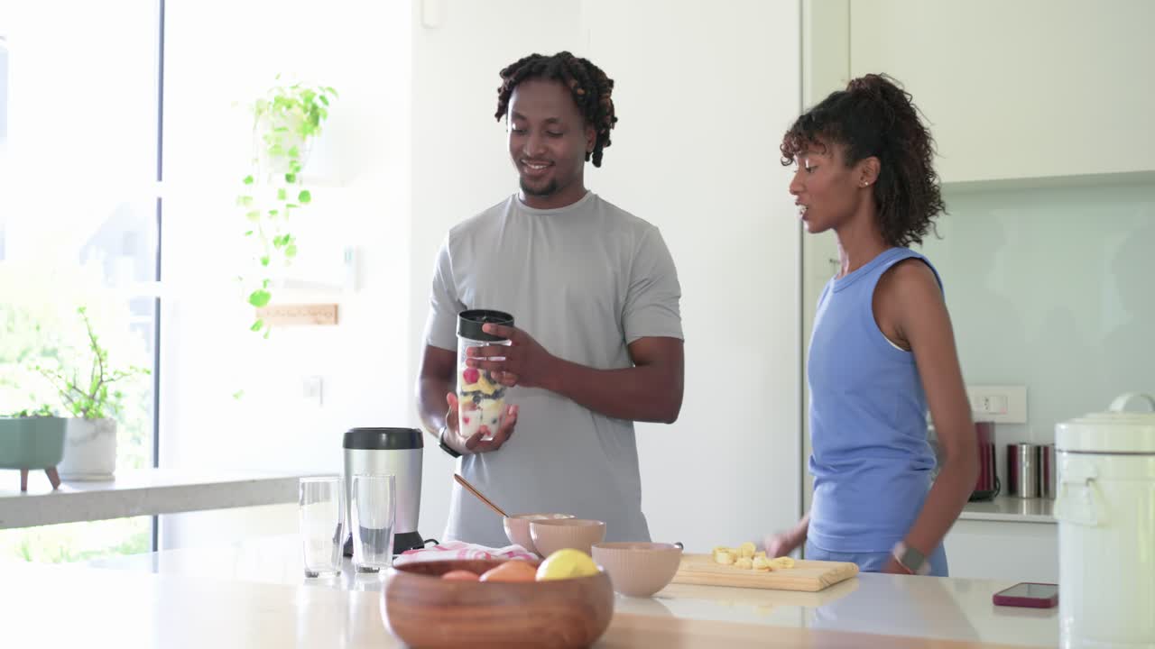 Coppia afroamericana che prepara un frullato in cucina; la donna gesticola mentre l'uomo, abbracciandola, riempie il bicchiere del frullatore.