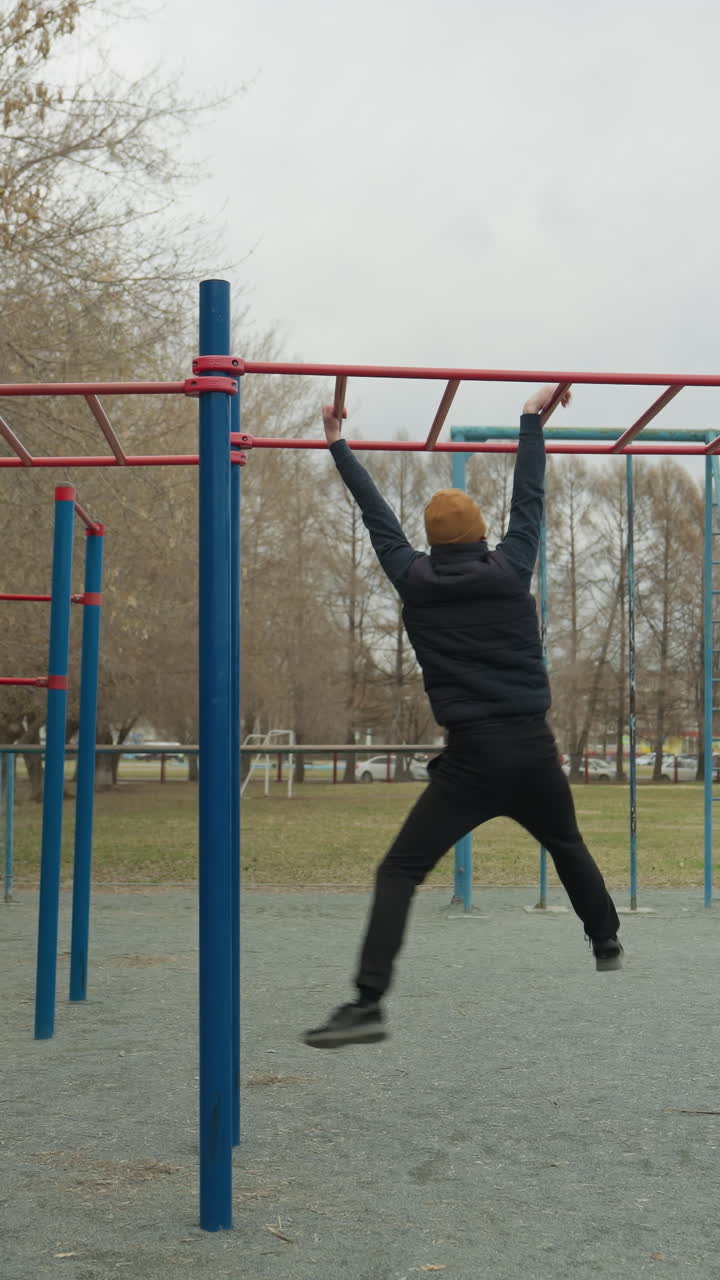 A coach leaps up to grab a red iron bar and steadily moves across it, after reaching the end of the bar, he drops down, in the background, a boy is playing football in a stadium