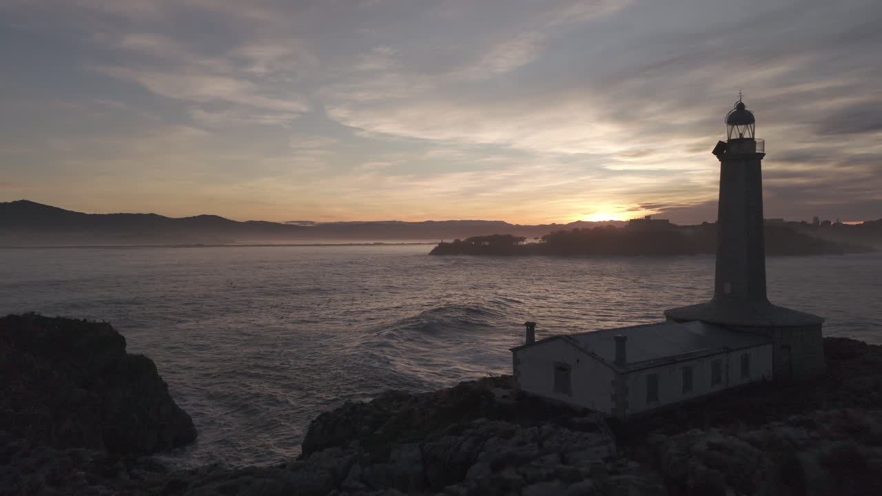 Tramonto drammatico su faro lungo una costa rocciosa con onde che si infrangono