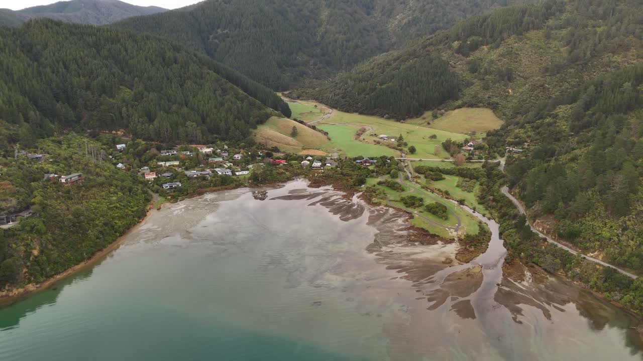 Whatamango Bay at low tide, coastal village nestled between lush green hills and water, near Picton, Marlborough Sounds, New Zealand. Aerial tilt-down forward