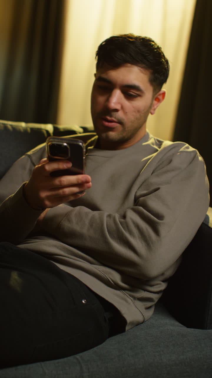 Vertical Video Of Young Man Spending Evening At Home Sitting On Sofa With Mobile Phone Scrolling Through Internet Or Social Media 1