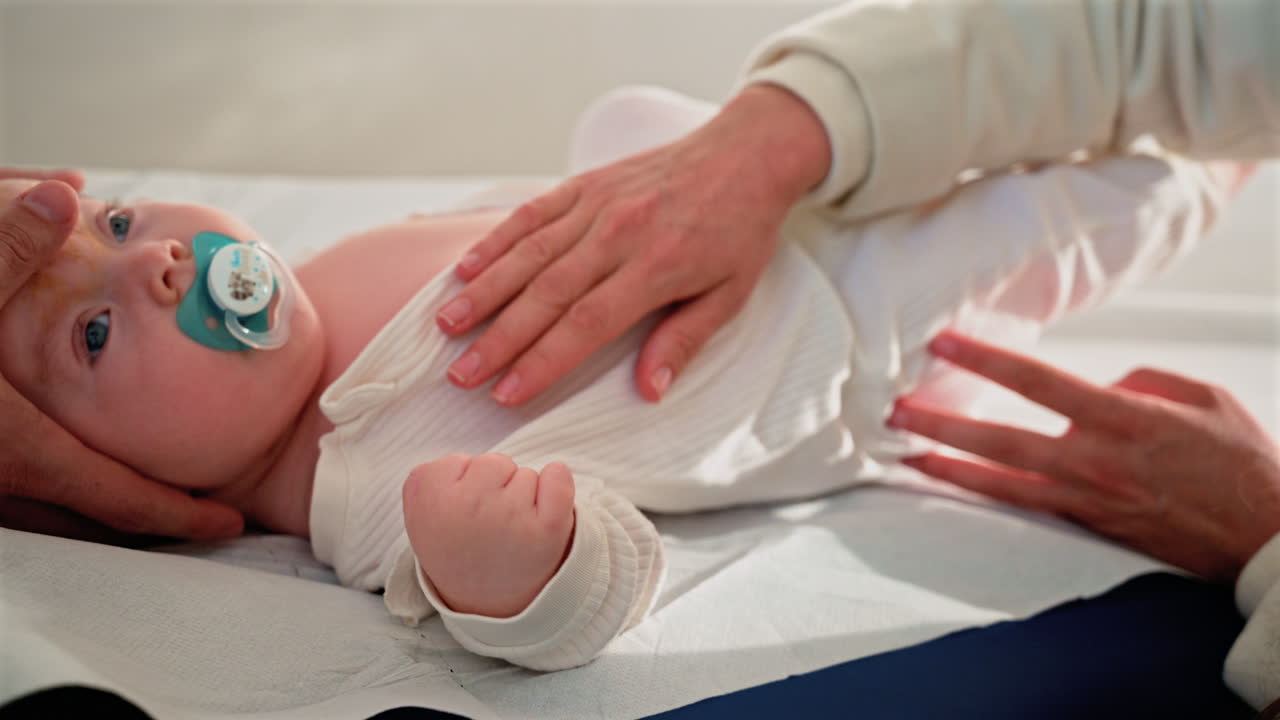 Primo piano di un neonato sdraiato su un lettino da visita medica, che tiene in mano un ciuccio mentre un adulto lo sostiene delicatamente