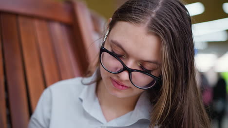 close-up of young adult with head slightly bent, wearing glasses, thoughtfully focusing on something in front of her, blurred background with unclear figures adds depth to the scene