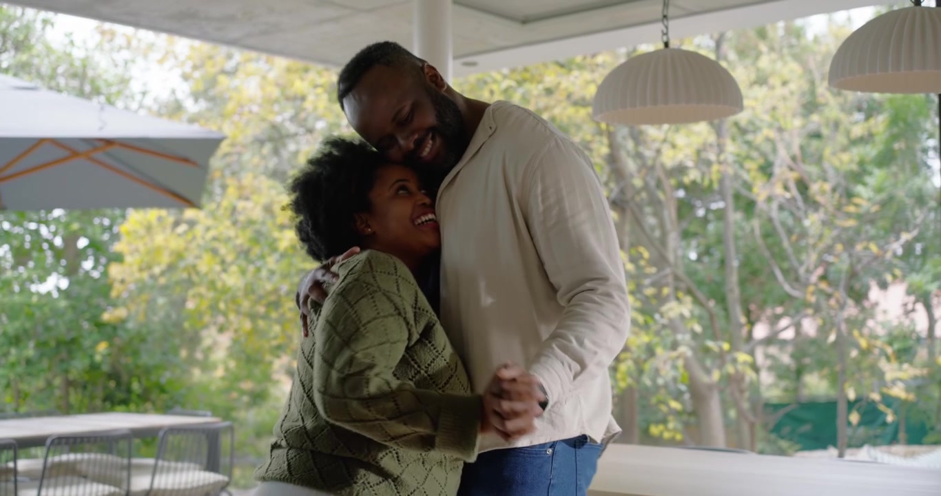 African American couple taking hands, dancing on covered patio with table, sharing affection