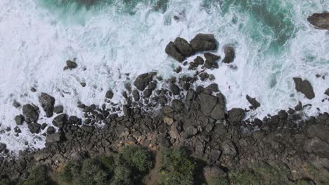top down drone shot of waves crashing rocky coastline in indonesia during sunny day