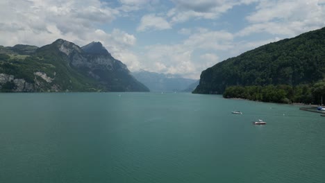 Barcos-Navegando-En-El-Lago-Frente-A-Un-Pequeño-Pueblo-En-Gäsi-Betlis,-Walensee-Glarus,-Weesen-Walenstadt,-Suiza--Drone-Shot