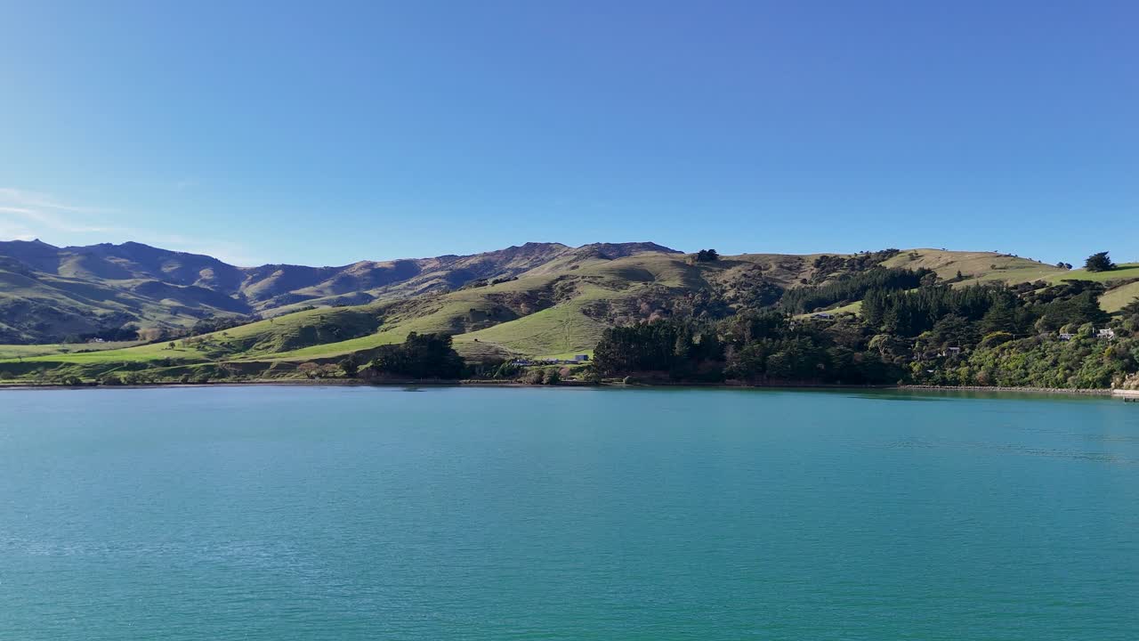 Aerial footage captures Akaroa's serene lake and rolling hills under clear blue skies, showcasing natural beauty and tranquility
