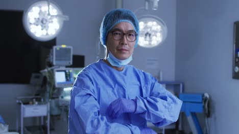 Portrait-of-asian-male-surgeon-wearing-lowered-face-mask,-crossing-arms-in-operating-theatre