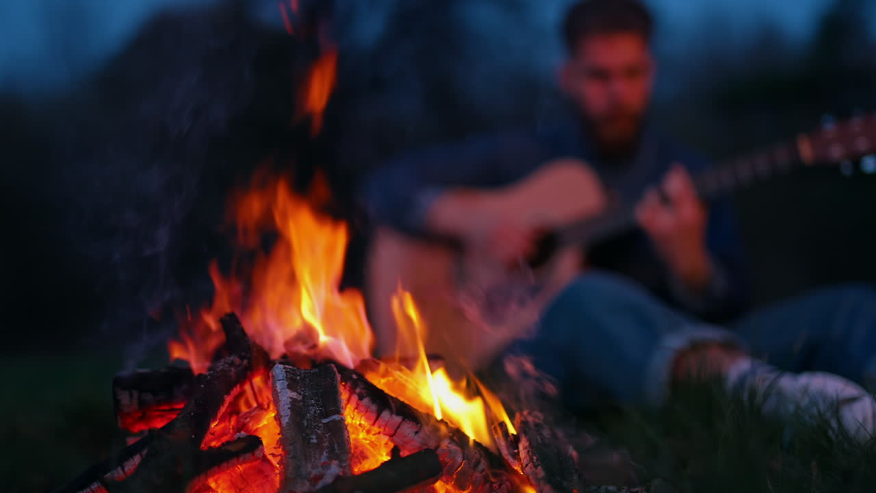 Beautiful campfire at night. Red and orange flame burning on blur background of a man playing the guitar in the evening. Leisure time outdoors.