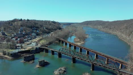 aerial establishing dolly of river town with train tracks on bridge, surrounded by mountains and blue skies in a scenic late fall or early spring landscape