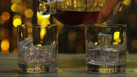 Pouring-of-whiskey,-cognac-or-brandy-from-bottle-into-glasses-with-ice-cubes.-Shiny-background