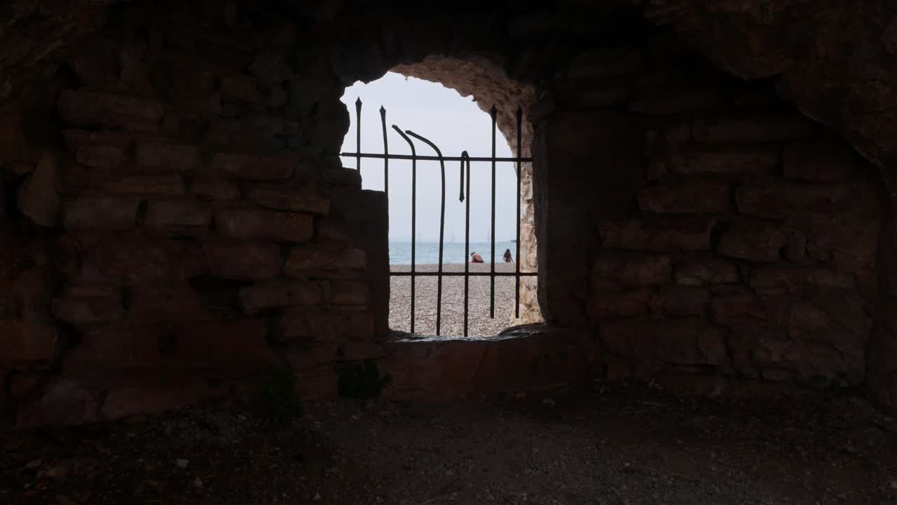 A Person Sits on the Beach Viewed Through a Ruined Stone Archway