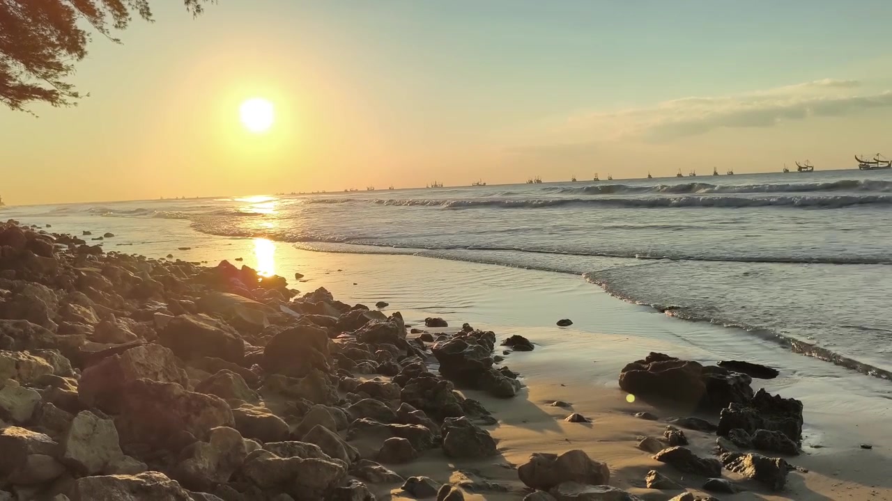 Pôr do Sol Dourado Sobre uma Praia Rochosa com Barcos de Pesca no Horizonte.