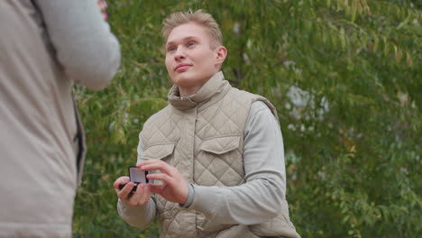 young man kneeling outdoors smiling warmly holding ring box proposing to woman with back slightly visible surrounded by swaying green leaves during romantic moment