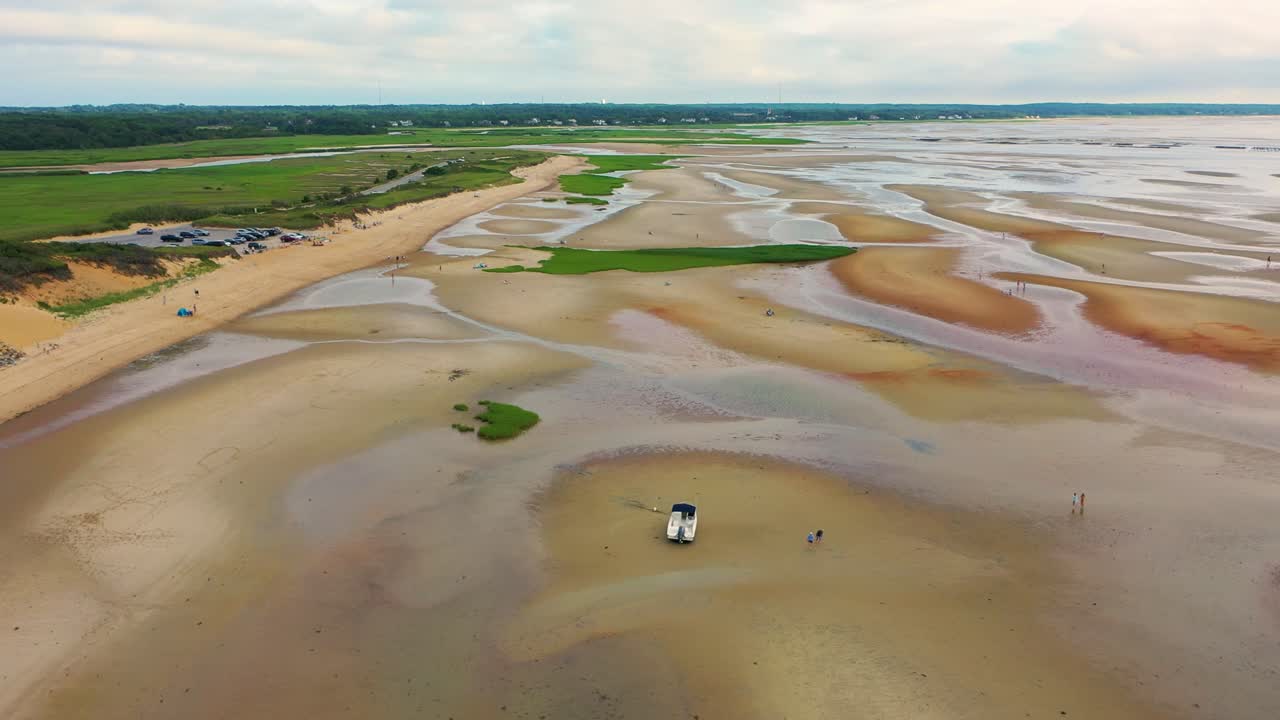 Drone footage captures a boat resting on wide tidal flats at low tide, with rippled sand patterns, shallow tide pools, and beachgoers walking along the exposed shoreline under cloudy skies