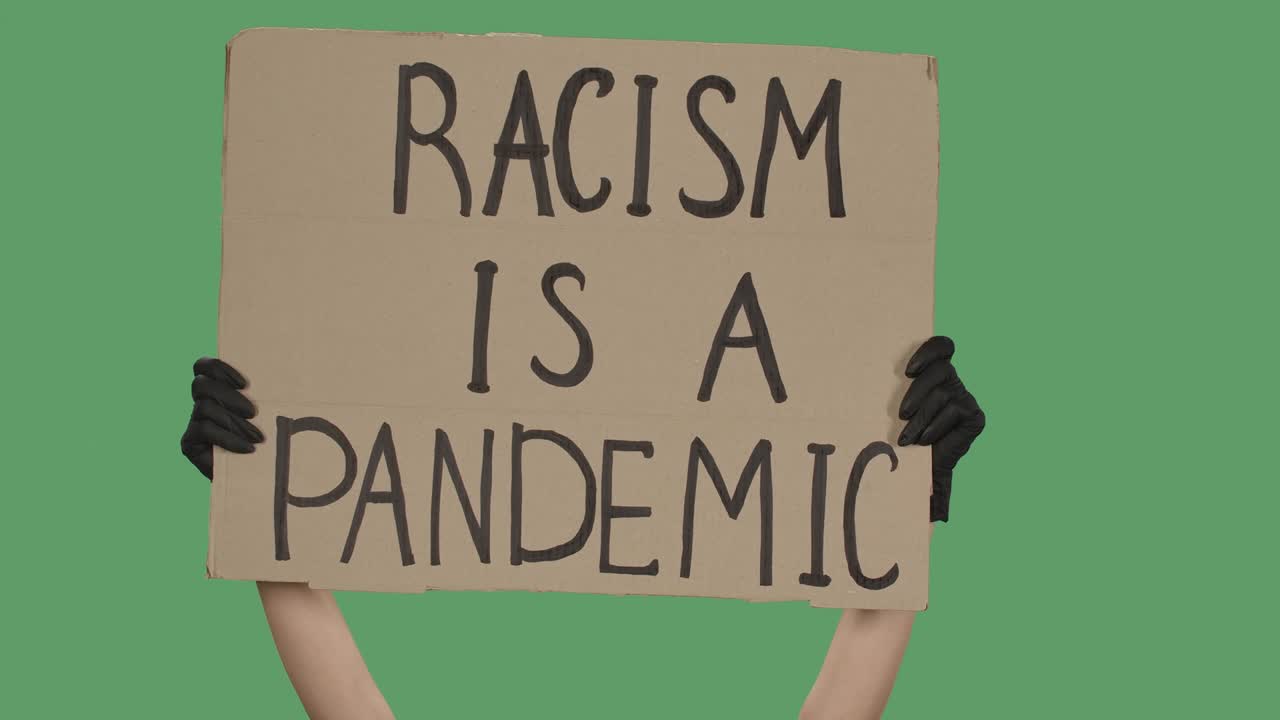 The hands of an unknown person in black gloves raise a cardboard poster RACISM IS A PANDEMIC. Equality and unity concept. Isolated a green screen, chroma key. Close up. Slow motion