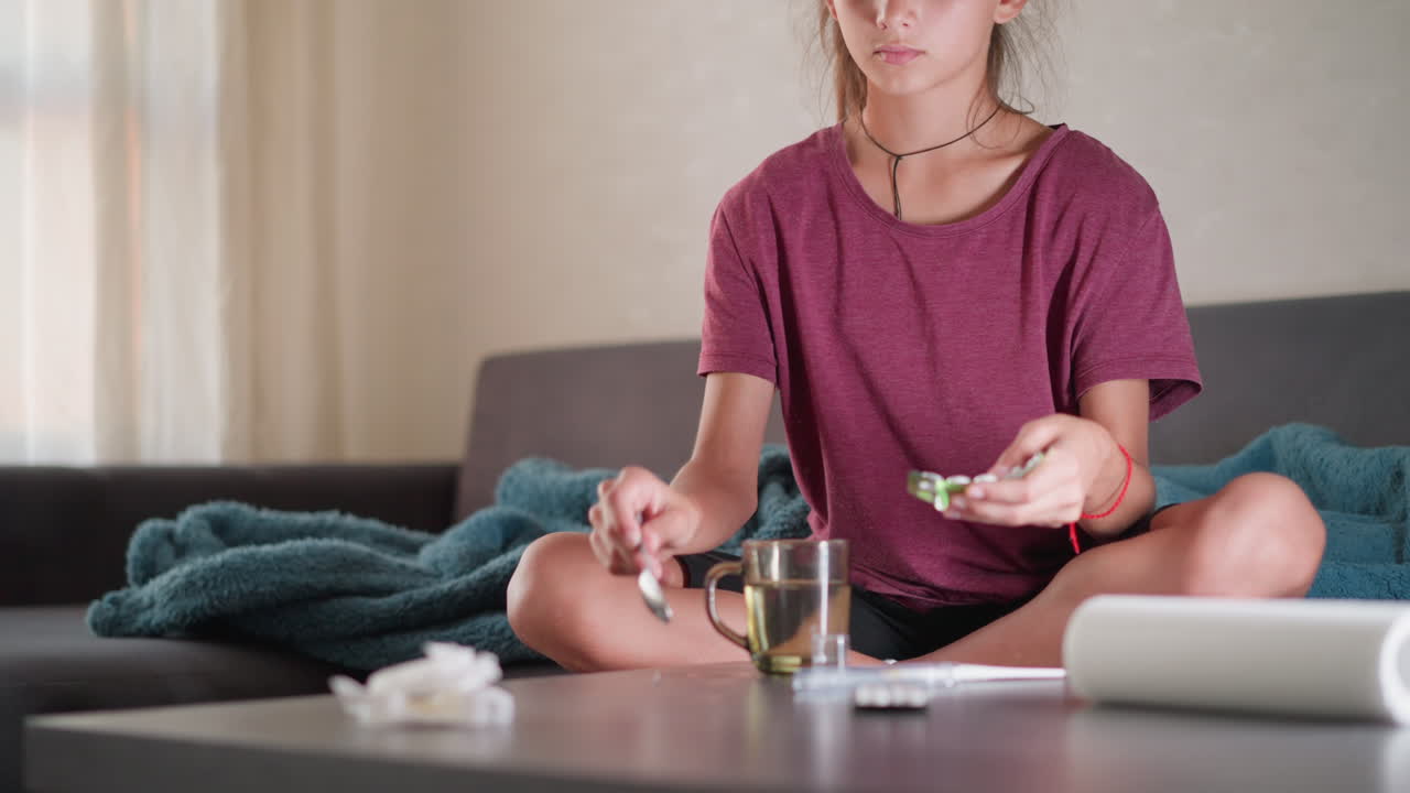 Young girl seated on couch prepares medicine in transparent cup using spoon while caring for sick brother lying nearby under blanket during home recovery with warm nurturing
