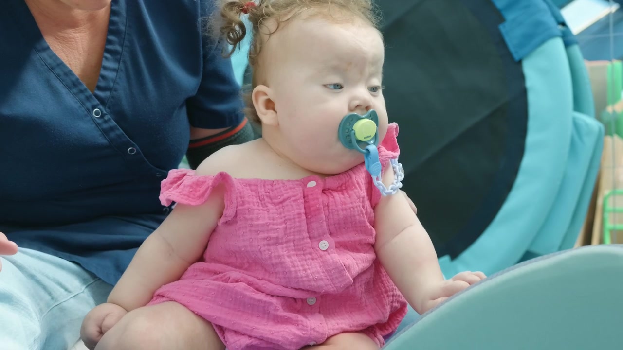 A baby girl sits on a mat with an adult nearby indoors