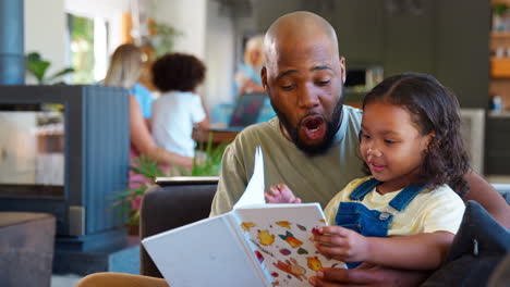 Father-And-Daughter-Reading-Book-At-Home-Together-With-Multi-Generation-Family-In-Background