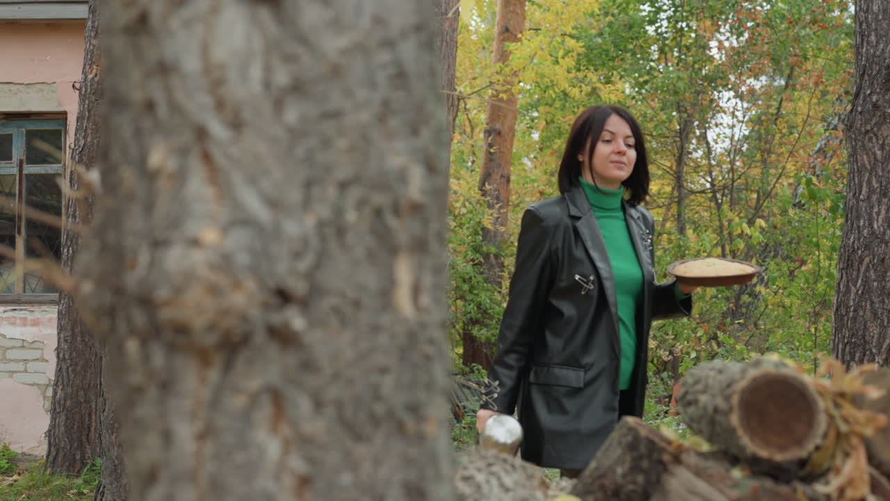 donna con giacca di pelle che esplora un cottage, radura nella foresta autunnale con foglie gialle, facciata rosa consumata e porta in legno, catasta di legna con pezzi appena tagliati, la donna posiziona una fetta di legno e poi versa