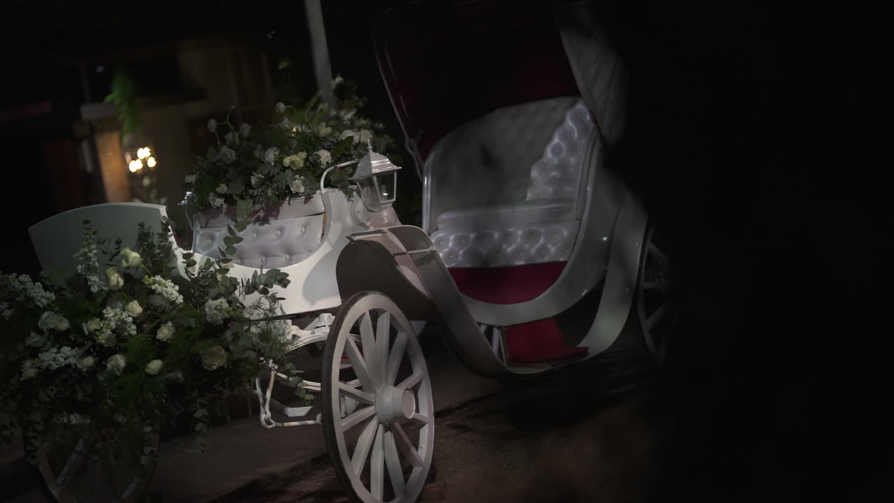 Romantic white horse-drawn carriage decorated with flowers and greenery, illuminated by a lantern, creating a magical atmosphere for a wedding celebration at night, slow motion reveal