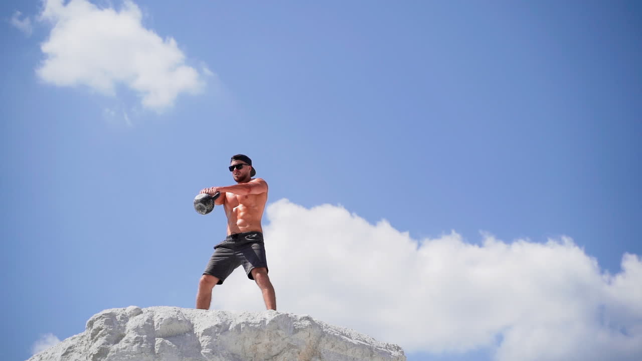 Athlete with kettlebell on white hill. Sportsman in black cap and sunglasses doing his workout on the mountain in a summer day. Slow motion
