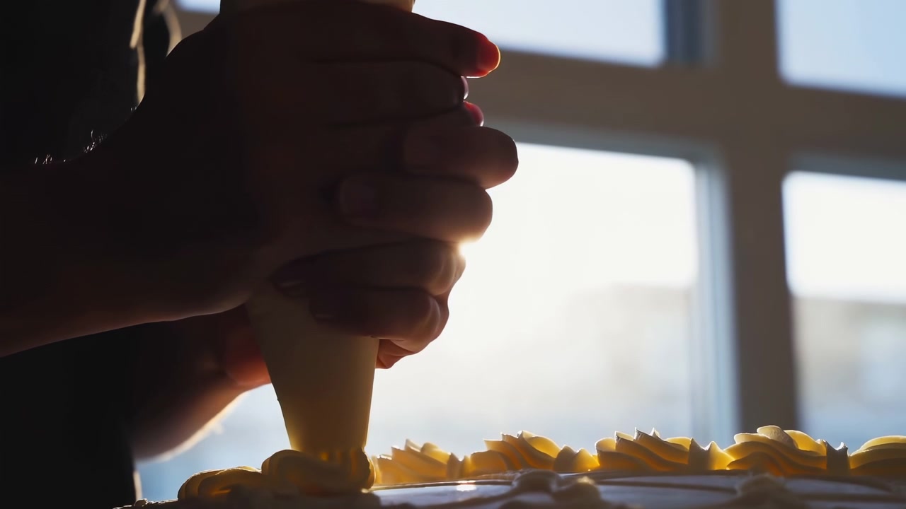 Close-up de mãos segurando um saco de confeitar e aplicando rosetas de creme de manteiga ao longo da borda do bolo enquanto a luz solar intensa entra por uma janela de vários painéis atrás do sujeito