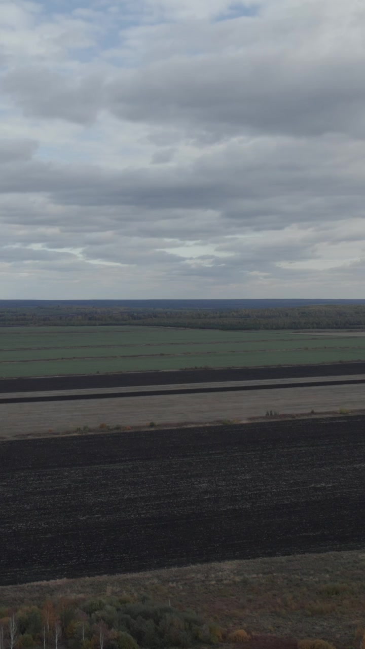 Drone footage of wide agricultural landscape with plowed soil stripes, green fields, and distant forest horizon under overcast clouds, rural countryside scenery
