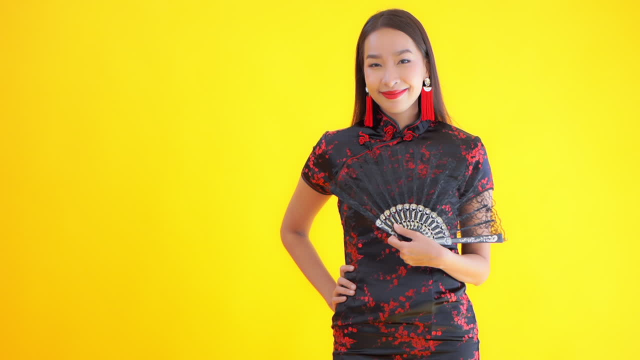 Asian woman in Chinese traditional costume dress holding the arm on the waist and fan oneself on studio yellow background