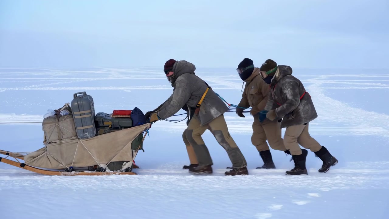 Three bundled people lean forward together as they push a gear-loaded sled across a flat snowy landscape under pale winter daylight, showing teamwork, travel