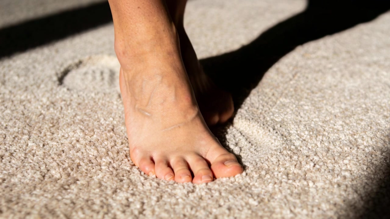 Walking barefoot person pressing ball after toe contact into plush carpet in room, with long shadow