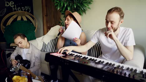 musical band is practising at home, guitarist is sitting on floor and playing the guitar, smiling vocalist is singing, keyboardist is laughing and playing the keyboard.