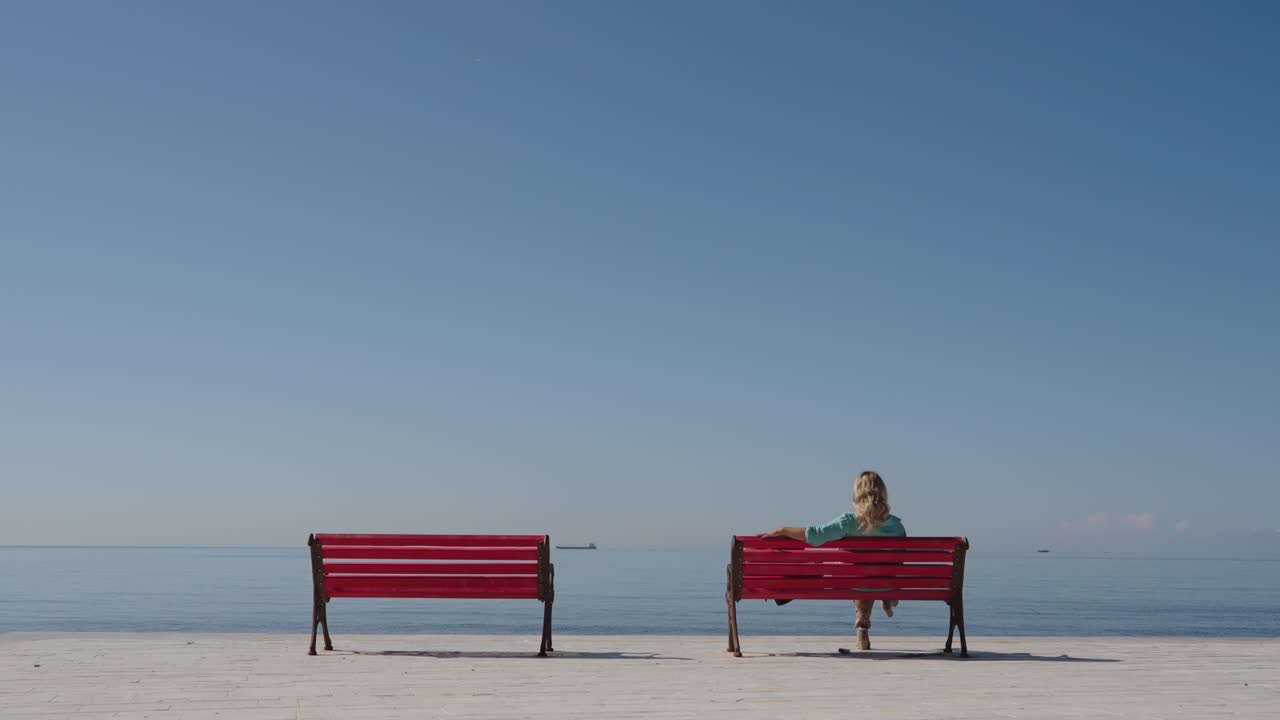 Blonde female sits alone on a red bench near a waterfront promenade, wide shot