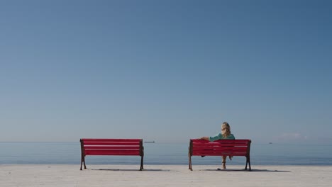blonde female sits alone on a red bench near a waterfront promenade, wide shot