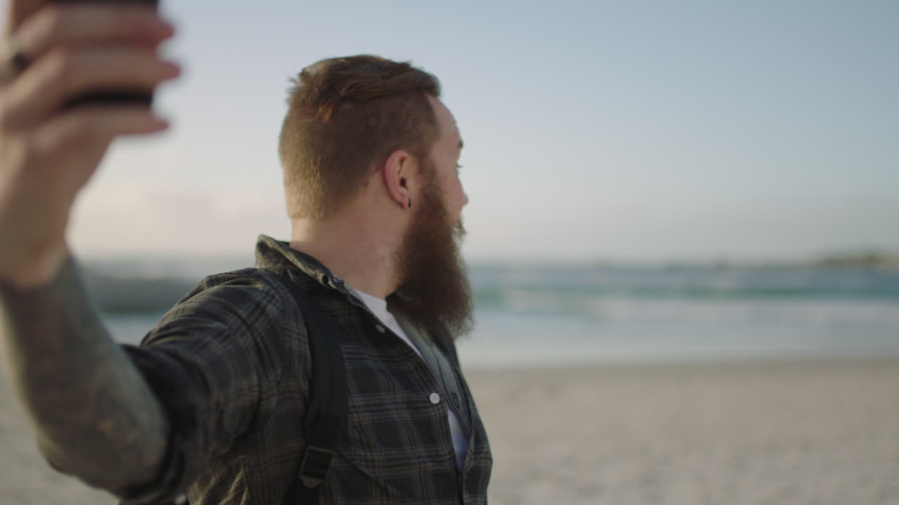 atractivo hombre hipster con barba usando teléfono para conversar por video en la playa hablando saludando