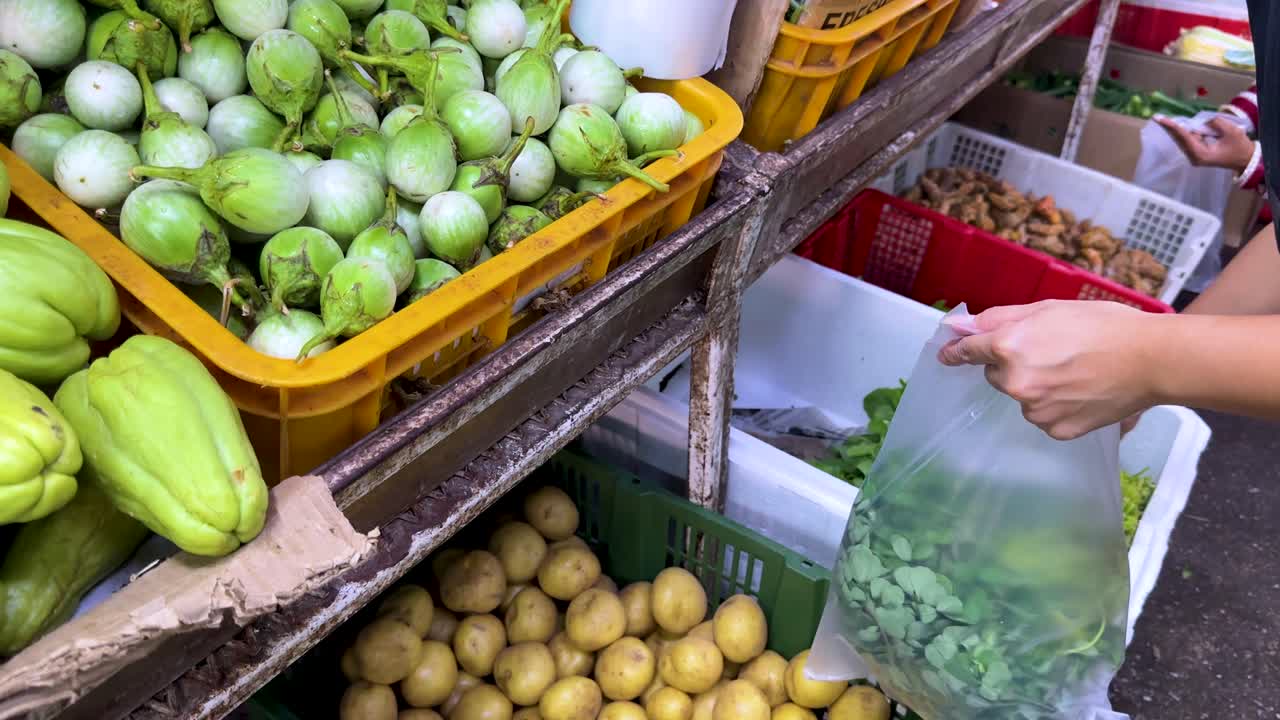 A person picks leafy greens and places them in a plastic bag at a vibrant outdoor vegetable stall, with natural daylight and handheld camera movement