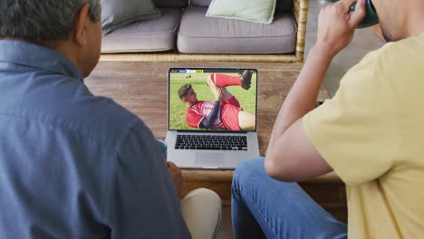 padre y hijo biraciales viendo una computadora portátil con varios jugadores de fútbol masculino jugando un partido en la pantalla