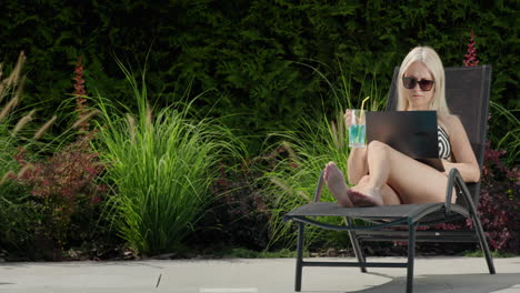 A-young-woman-in-a-swimsuit-is-relaxing-on-a-sun-lounger,-using-a-laptop.-Holds-a-cool-cocktail-with-ice-in-his-hand